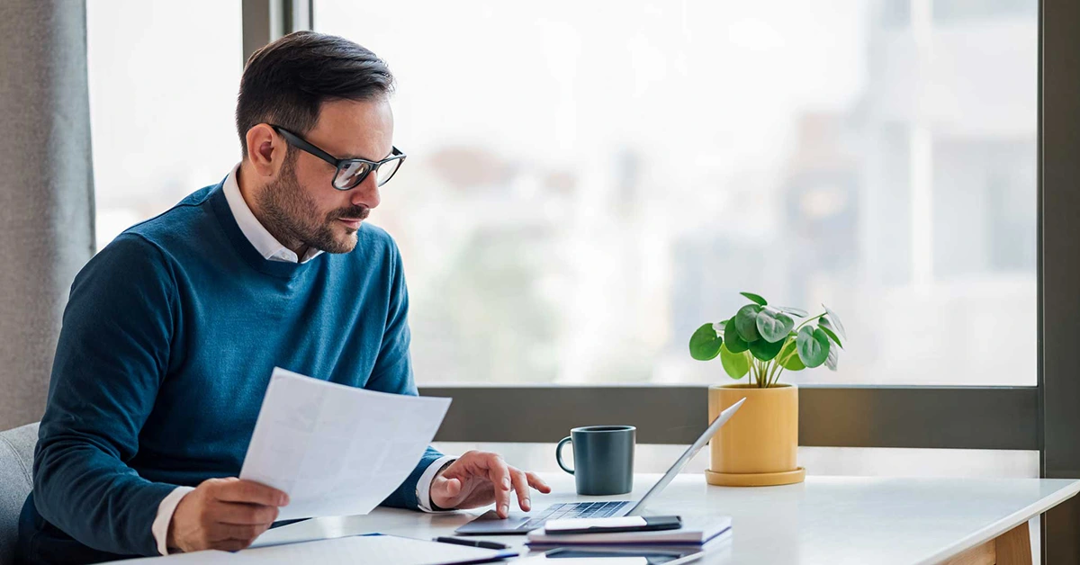 A business man sits by a commercial window, comfortably, while working on his computer.
