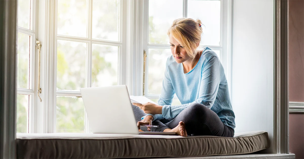 A woman sits by her window, comfortably, while paying bills on her computer.