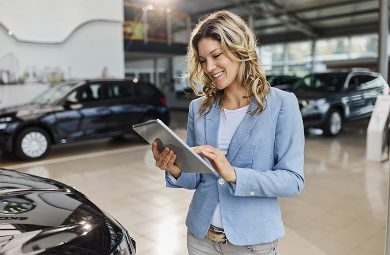 A sales woman looks a tablet comfortably inside a car dealership with large bay windows.