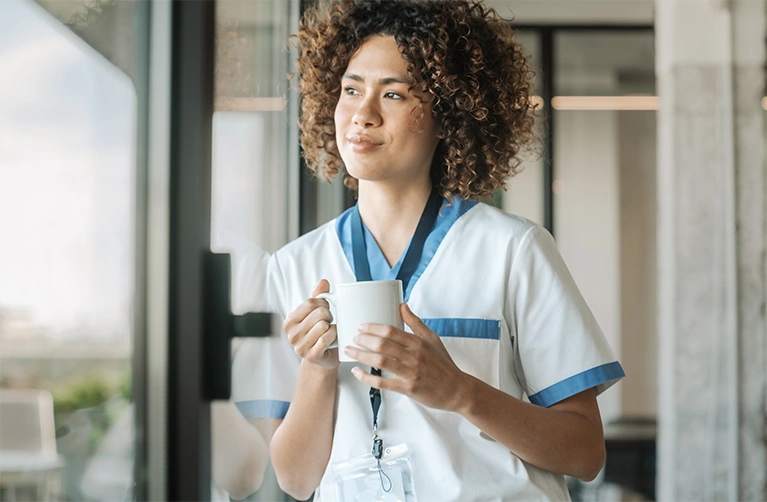 A woman wearing medical scrubs and holding a coffee cup looks out a window from a medical office.
