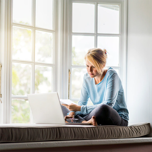 A woman sits by her window, covered with window film, looking at her laptop comfortably.