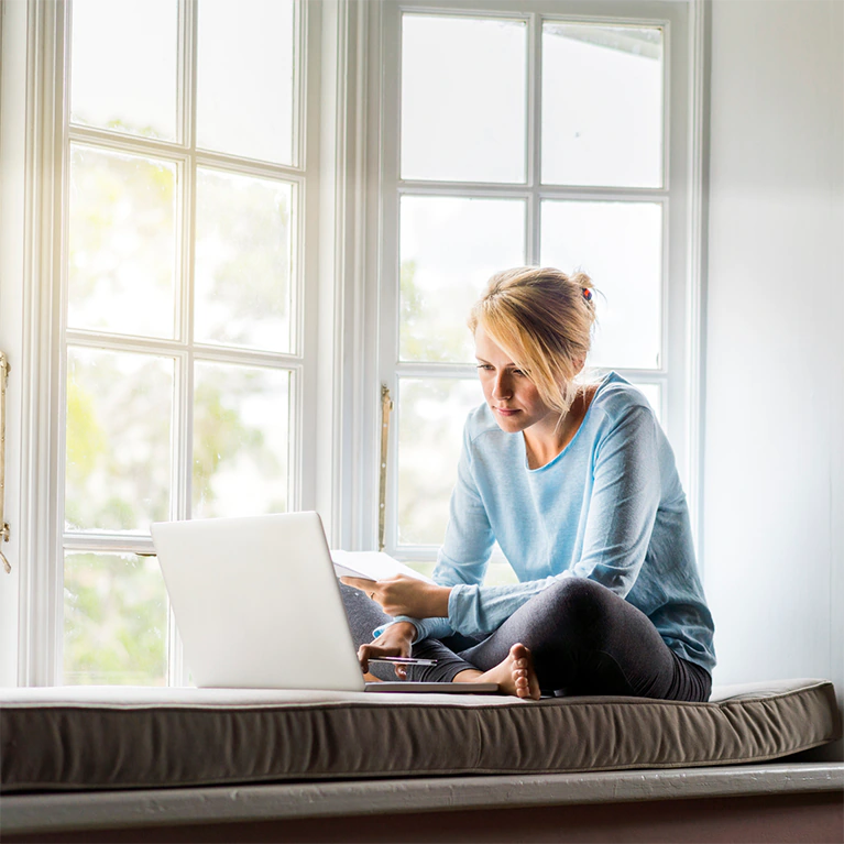 A woman sits by her window, covered with window film, looking at her laptop comfortably.