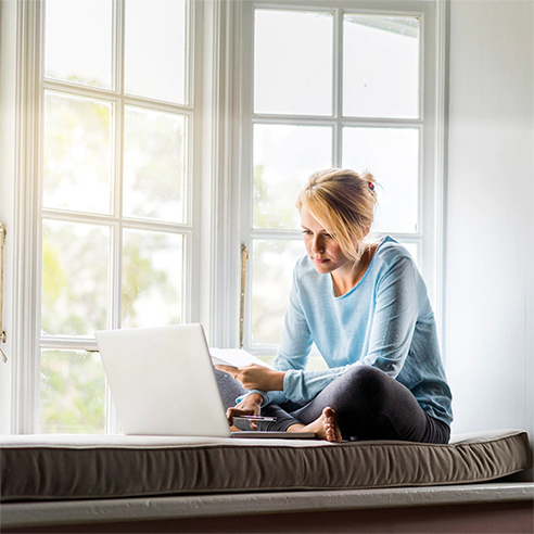 A woman sits by her window, covered with window film, looking at her laptop comfortably.