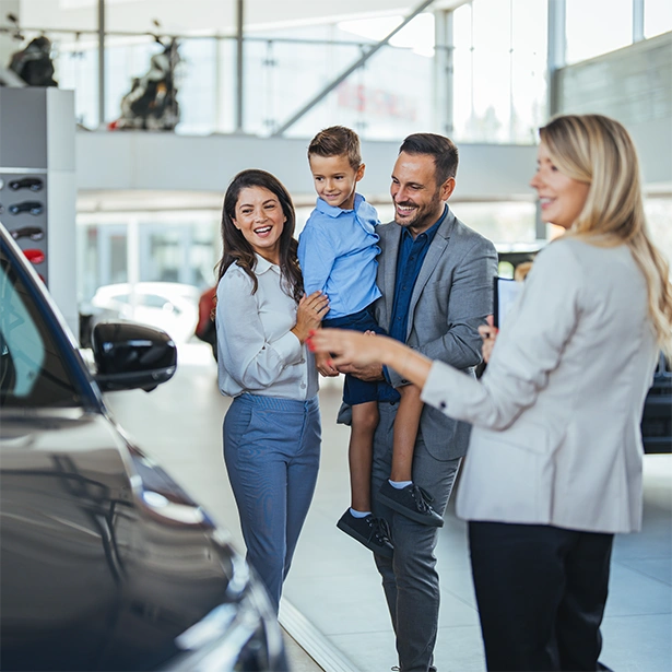 A sales woman shows a family a car at a dealership with large bay windows.