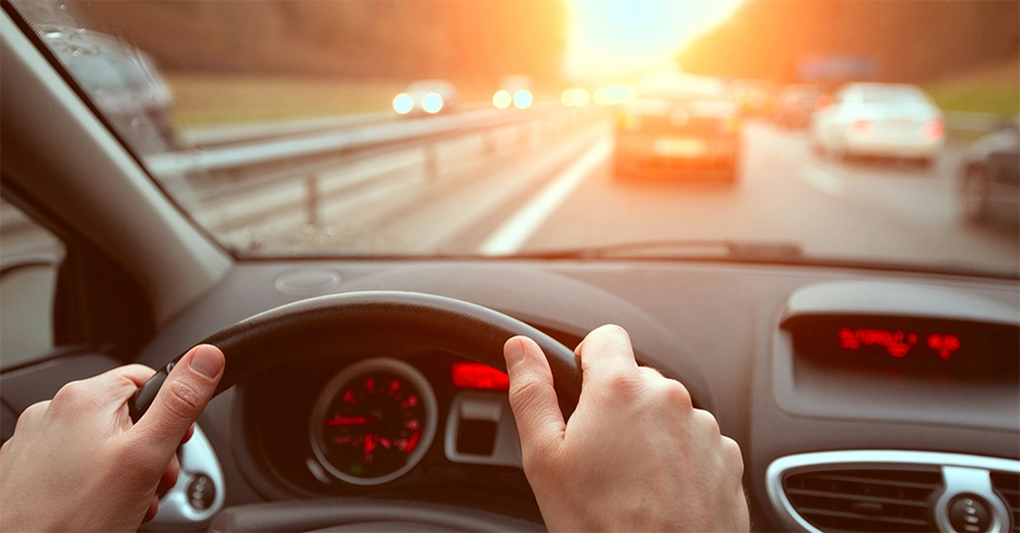 Hands are on a steering wheel as someone drives down a highway toward glare.