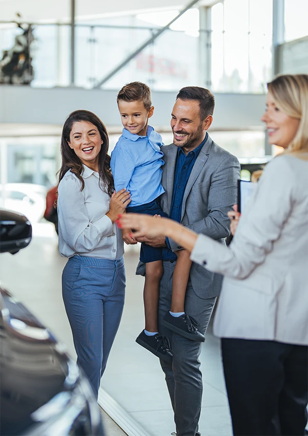 A saleswoman shows a family of three a car at a dealership with large bay windows behind them.